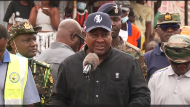 A man speaks into a microphone outdoors while surrounded by uniformed personnel and other attendees.