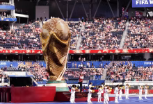 A large World Cup trophy display stands on a stadium field while performers in white line up in front of a seated crowd.