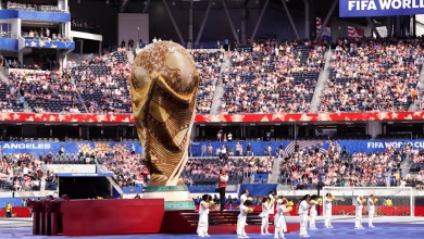 A large World Cup trophy display stands on a stadium field while performers in white line up in front of a seated crowd.