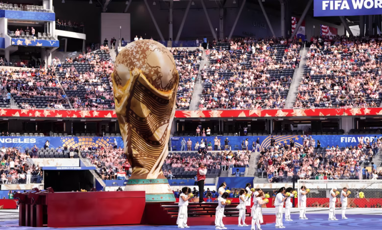 A large World Cup trophy display stands on a stadium field while performers in white line up in front of a seated crowd.