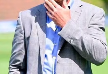A cropped outdoor image shows a person in a light gray suit jacket and blue shirt holding a hand near their face on a grassy field.