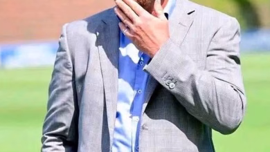 A cropped outdoor image shows a person in a light gray suit jacket and blue shirt holding a hand near their face on a grassy field.