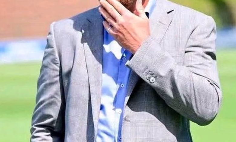 A cropped outdoor image shows a person in a light gray suit jacket and blue shirt holding a hand near their face on a grassy field.