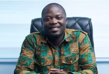 A man wearing a patterned shirt sits smiling in an office chair against a light background.