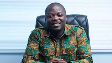 A man wearing a patterned shirt sits smiling in an office chair against a light background.