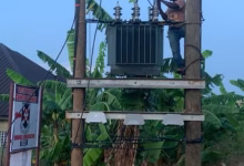 A worker stands on a utility pole beside an electrical transformer in a leafy residential area.
