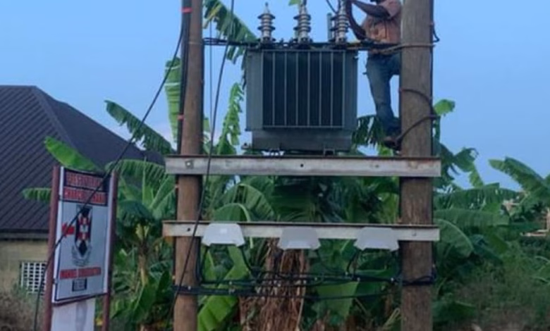 A worker stands on a utility pole beside an electrical transformer in a leafy residential area.