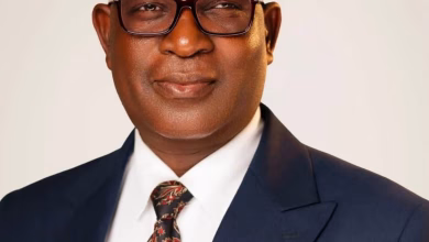 A cropped formal portrait shows a man wearing glasses, a dark suit, white shirt, and patterned tie against a plain light background.