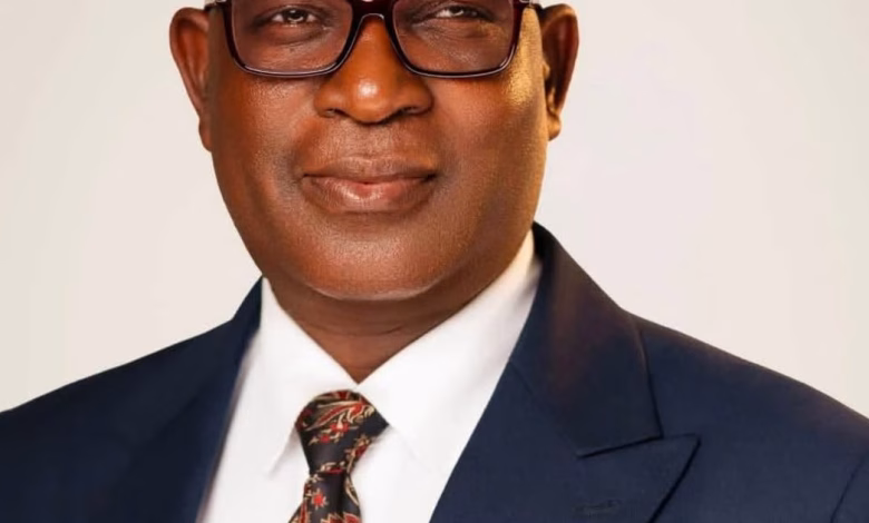 A cropped formal portrait shows a man wearing glasses, a dark suit, white shirt, and patterned tie against a plain light background.