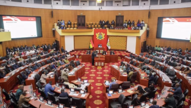 A wide view shows legislators seated in a large parliamentary chamber with a red carpeted central aisle and a speaker at the front.