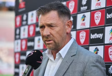 A man in a grey suit is being interviewed with a microphone in front of a sports sponsor backdrop.