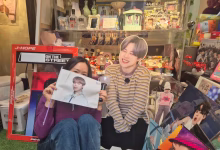 A person sits in front of a densely decorated display of pop-music memorabilia while holding a portrait and posing beside a life-size cardboard cutout.