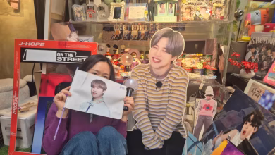 A person sits in front of a densely decorated display of pop-music memorabilia while holding a portrait and posing beside a life-size cardboard cutout.