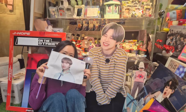 A person sits in front of a densely decorated display of pop-music memorabilia while holding a portrait and posing beside a life-size cardboard cutout.