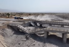 A damaged concrete bridge spans a dry, rocky landscape, with a truck on a nearby road and dust rising in the background.