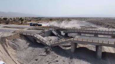 A damaged concrete bridge spans a dry, rocky landscape, with a truck on a nearby road and dust rising in the background.
