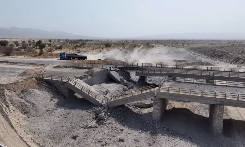 A damaged concrete bridge spans a dry, rocky landscape, with a truck on a nearby road and dust rising in the background.