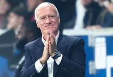 An older man in a dark suit applauds while seated in a stadium or event setting with a blurred crowd behind him.