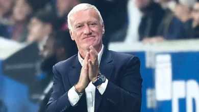 An older man in a dark suit applauds while seated in a stadium or event setting with a blurred crowd behind him.