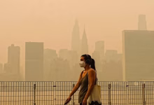 A person wearing a face mask walks beside a metal fence with a hazy city skyline in the background.