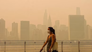 A person wearing a face mask walks beside a metal fence with a hazy city skyline in the background.