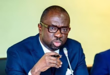 A man in a dark blue suit and glasses speaks into a tabletop microphone against a pale yellow-green background.