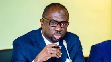 A man in a dark blue suit and glasses speaks into a tabletop microphone against a pale yellow-green background.