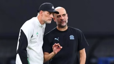 Two men in sportswear stand together on the sidelines, with one appearing to gesture while speaking.