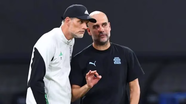 Two men in sportswear stand together on the sidelines, with one appearing to gesture while speaking.