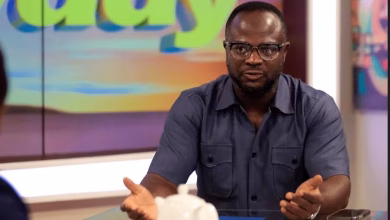 A man wearing glasses and a dark short-sleeved shirt gestures while seated at a studio table, with a colorful screen behind him.