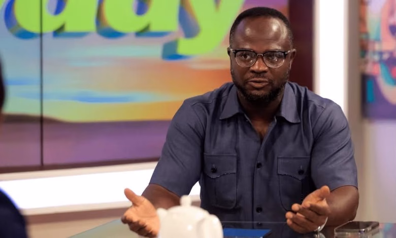 A man wearing glasses and a dark short-sleeved shirt gestures while seated at a studio table, with a colorful screen behind him.