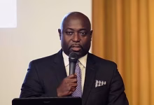 A man in a dark suit speaks into a handheld microphone while standing behind a laptop indoors.