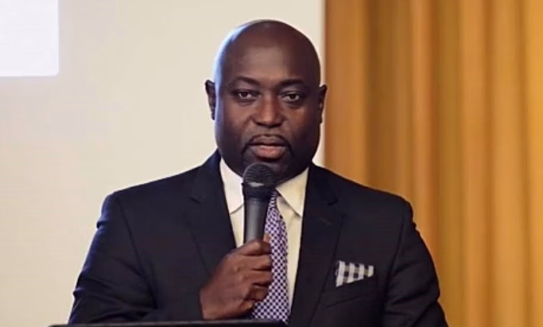 A man in a dark suit speaks into a handheld microphone while standing behind a laptop indoors.