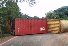 Two shipping containers lie overturned across a paved road bordered by dense trees and vegetation.