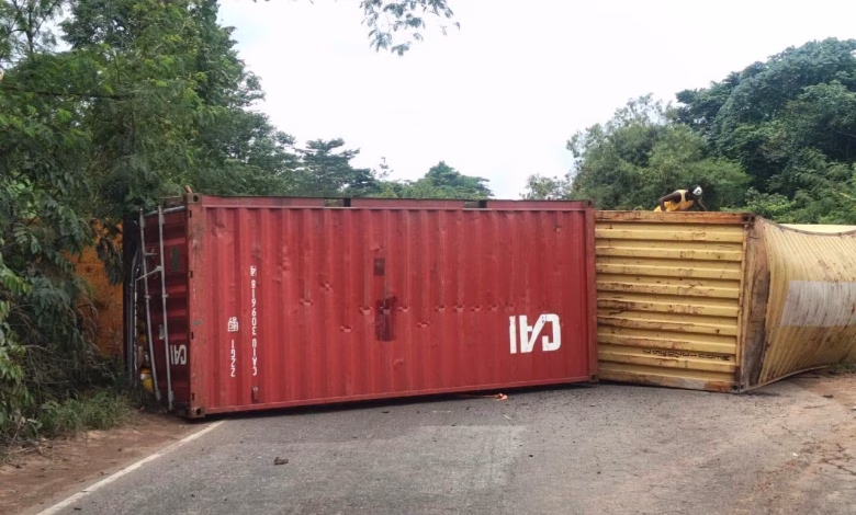 Two shipping containers lie overturned across a paved road bordered by dense trees and vegetation.