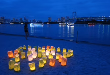 Illuminated paper lanterns sit on a beach at dusk beside calm water, with a bridge and city skyline in the background.