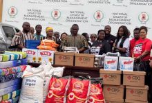 A group of people stands behind stacks of donated household supplies and boxed goods in front of a National Disaster Management Organisation backdrop.